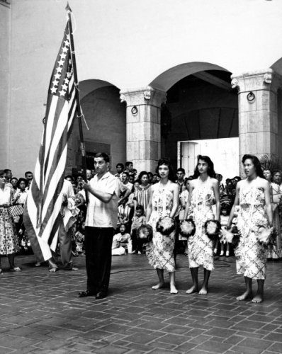 Man holding the flag of the United States with temporary stars accompanied by hula dancers at Honolulu Hale (City Hall) in celebration of statehood; Honolulu, Hawai‘i, March 12, 1959. Photograph by Irving Rosen. Call Number: Rosen Collection; Unp. Image from Bishop Museum Archives, Honolulu, Hawaii. Images are not to be re-used without permission.