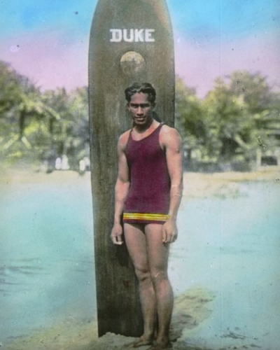 Duke Kahanamoku standing in front of his surfboard; Waikīkī, Hawai‘i. ca. 1915. Hand-tinted lantern slide. Collection: General Photograph Collection Call Number: People. Kahanamoku, folder 2. Artifact Number: SLS 24263 Location: Bishop Museum Archives Image from Bishop Museum Archives, Honolulu, Hawaii. Images are not to be re-used without permission.