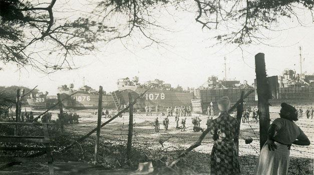 Just after the Dec. 7th attack, barbed wire was hurriedly strung along many miles of Hawaiian coastlines and around important buildings. Beachside residents found themselves cut off from access to the ocean. In some locations, access was even more restricted by military maneuvers like what is shown in this photo taken in Kīhei, Maui. Landing craft line the beach as men practice coming ashore, in preparation for fighting to retake Pacific islands held by the Japanese. Meanwhile, two women watch from a home’s front yard. Collection: General Photograph Collection Call Number: Military. War. WWII. Amphibious Training. Artifact Number: SP 96137 Location: Bishop Museum Archives Image from Bishop Museum Archives, Honolulu, Hawaii. Images are not to be re-used without permission.