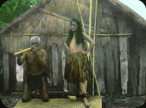 Man playing the nose flute and hula dancer in front of hale pili (grass house), Hawai‘i. ca. 1890-1905. Hand-colored lantern slide. Photographer: Henry W. Henshaw. Call Number: E.C. Hawaiian. Music. Instruments. Image from Bishop Museum Archives, Honolulu, Hawaii. Images are not to be re-used without permission.